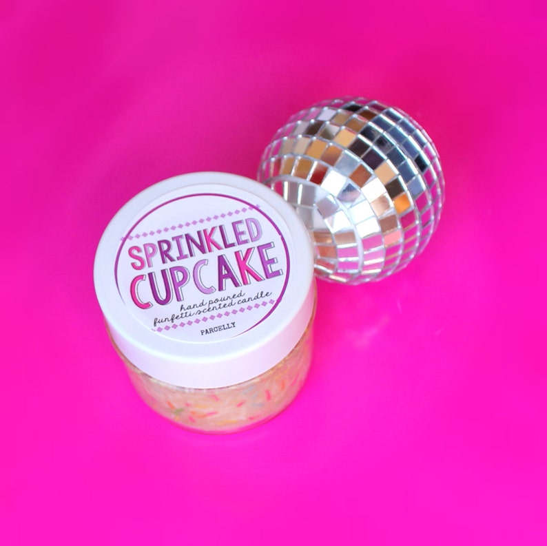 May include: A white jar with a pink and purple label that reads "SPRINKLED CUPCAKE hand poured confetti-scented candle PARCELLY". The jar is filled with a white substance with colorful sprinkles. A silver disco ball is in the background.