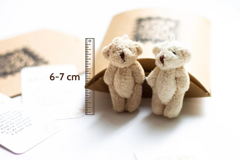 May include: Two small, beige teddy bears, each measuring 6-7 cm tall, are sitting on a brown paper bag. The bears have brown eyes and noses.