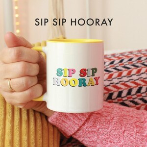 May include: A white ceramic mug with a yellow rim and handle. The mug has the text "Sip Sip Hooray" in colorful letters.