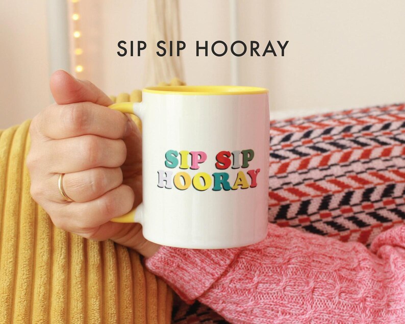 May include: A white ceramic mug with a yellow rim and handle. The mug has the text "Sip Sip Hooray" in colorful letters.