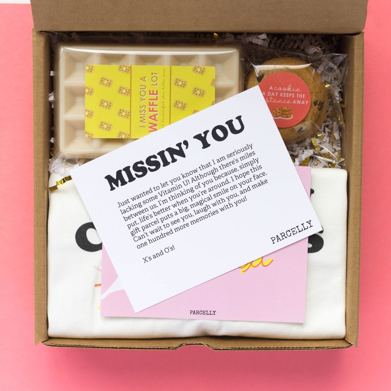 May include: A pink and white gift box with a white t-shirt, a yellow and white card with the text "Missin' You", a white waffle candle, a small bag of chocolate chips, and a small card with the text "A cookie a day keeps the distance away".
