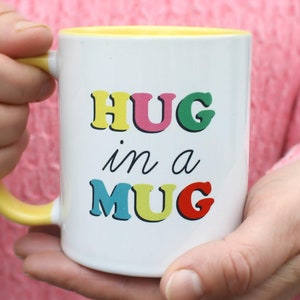 May include: White ceramic mug with a yellow rim and handle. The mug has the text "HUG in a MUG" printed on it in colorful letters.