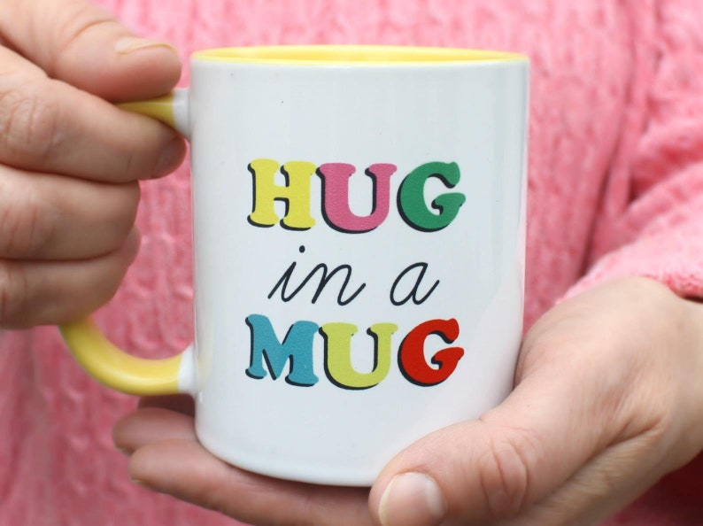 May include: White ceramic mug with a yellow rim and handle. The mug has the text "HUG in a MUG" printed on it in colorful letters.
