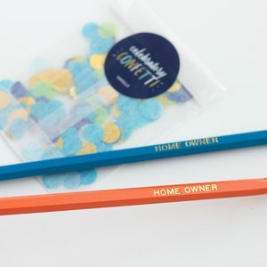 May include: Two pencils with gold lettering that reads "HOME OWNER" on a white background. One pencil is blue and the other is orange. A small bag of confetti is in the background.