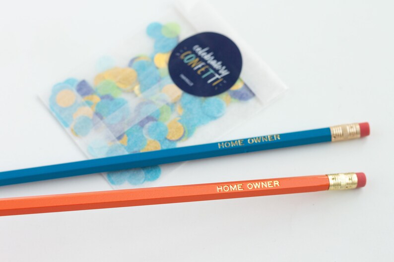 May include: Two pencils with gold lettering that reads "HOME OWNER" on a white background. One pencil is blue and the other is orange. A small bag of confetti is in the background.