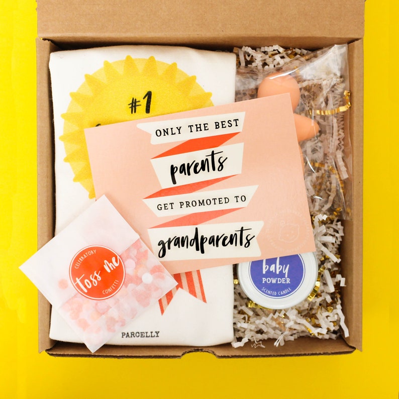 May include: A gift box filled with items for new grandparents. The box contains a white t-shirt with a yellow sun and the text "#1 ONLY THE BEST parents GET PROMOTED TO grandparents", a pink card with the text "ONLY THE BEST parents GET PROMOTED TO grandparents", a small bag of confetti, a small jar of baby powder scented candle, and a small pink baby doll.