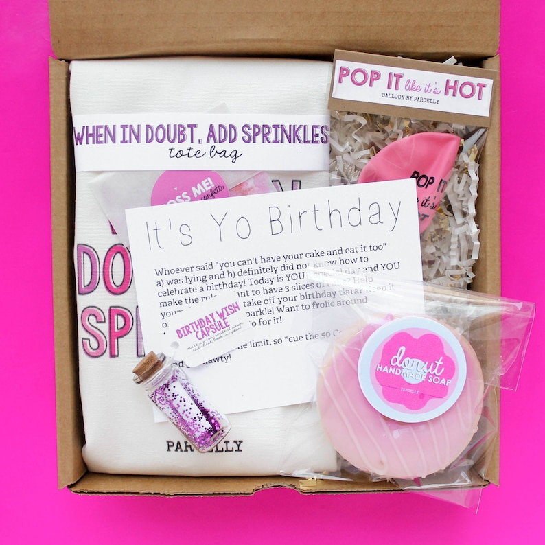 May include: A pink and white birthday gift box with a white tote bag that says "When in doubt, add sprinkles", a pink balloon, a pink donut soap, a small bottle of glitter, and a card that says "It's Yo Birthday".