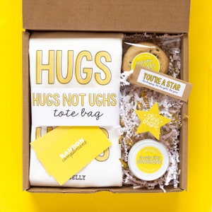 May include: A gift box filled with yellow and white themed items. The box contains a tote bag with the text "Hugs Not Ughs Tote Bag", a cookie, a candle, and a card that says "You're a Star, be happy be bright be you".