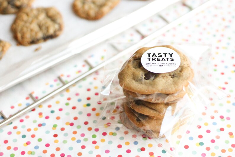 May include: A stack of four chocolate chip cookies in a clear plastic bag with a white circle sticker that says "Tasty Treats Chocolate Chip Cookies Made with Love" on a white surface with colorful polka dots.