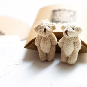 May include: Two small, plush teddy bears, one beige and one white, are sitting side-by-side on a brown paper surface. The bears have brown eyes and black noses.