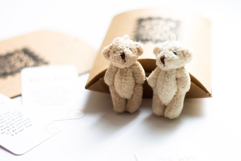 May include: Two small, plush teddy bears, one beige and one white, are sitting side-by-side on a brown paper surface. The bears have brown eyes and black noses.