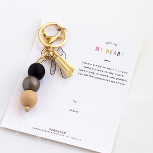 May include: A gold-toned keychain with a black, gray, and tan beaded design and a tassel. The keychain is displayed with a card that reads "KEY TO MY HEART" and includes a message about love and appreciation.