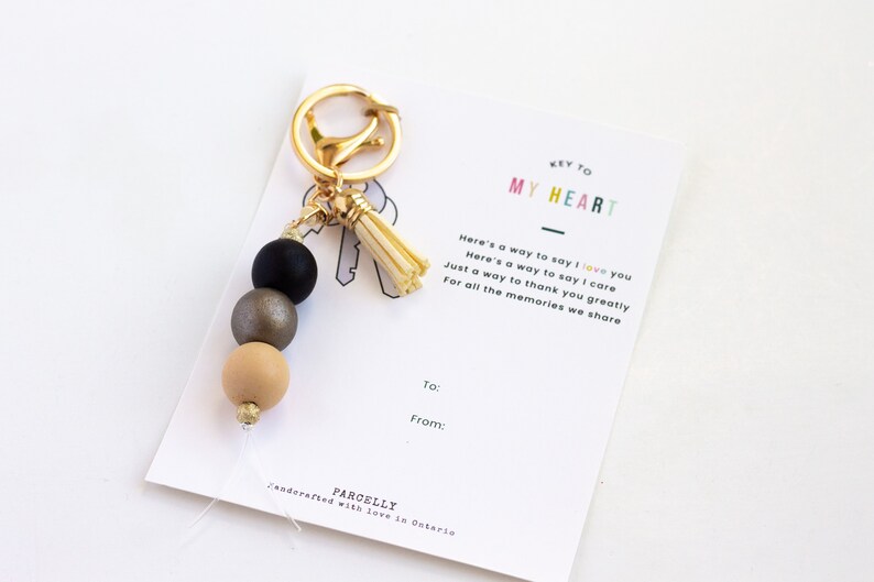 May include: A gold-toned keychain with a black, gray, and tan beaded design and a tassel. The keychain is displayed with a card that reads "KEY TO MY HEART" and includes a message about love and appreciation.