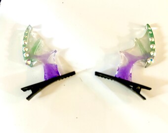 May include: A pair of purple and green iridescent hair clips with rhinestones. Each clip has a wing-like shape and is attached to a black alligator clip.