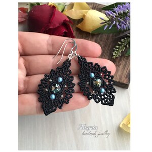 May include: Black dangle earrings with a delicate, intricate design. The earrings feature a black cord woven into a diamond shape with a blue crystal and pearl accents. The earrings are suspended from silver hooks.