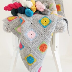 Può includere: Una coperta di uncinetto grigia con cerchi colorati, che presenta una varietà di colori di filato, tra cui rosa, blu, giallo, verde e arancione. La coperta è drappeggiata su una sedia bianca.