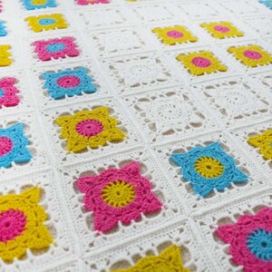 Può includere: Una coperta all'uncinetto bianca con quadrati colorati. Ogni quadrato presenta una diversa combinazione di colori rosa, giallo e blu.