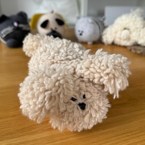 Op de afbeelding: Een pluche wit gehaakt hondenspeelgoed met zwarte ogen en een zwarte neus. Het speelgoed ligt op zijn rug met zijn poten in de lucht.