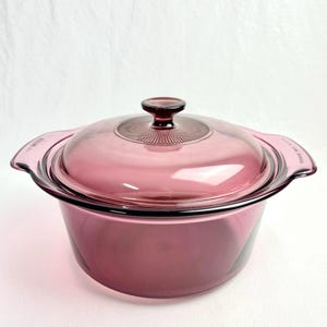 May include: A vintage, round, transparent, purple glass casserole dish with a matching lid. The lid has a knob handle and a ridged design. The dish has two handles. The cookware is designed for oven use.