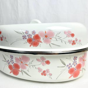 May include: White enamel casserole dish with a lid, decorated with pink, red, and purple floral patterns. The flowers are set against a white backdrop with grey stems and leaves. It has a silver rim and a handle on the lid. The dish is suitable for cooking.