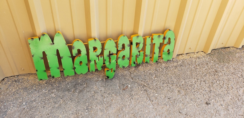May include: Green metal sign with the word "Margarita" in yellow outlined letters. The sign has a distressed look with black paint showing through the green paint.