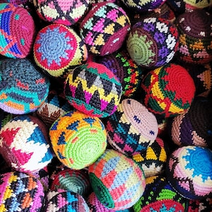 May include: A collection of colorful, hand-crocheted balls. Each ball features a unique pattern and color combination, including shades of red, blue, green, yellow, and purple. The balls appear to be made of yarn and are likely used for juggling or play.