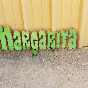 Puede incluir: Letras de metal verde deletreando la palabra "Margarita" sobre un fondo marrón claro.