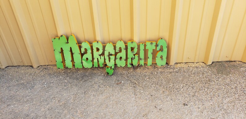 May include: Green metal letters spelling out the word "Margarita" on a light brown background.