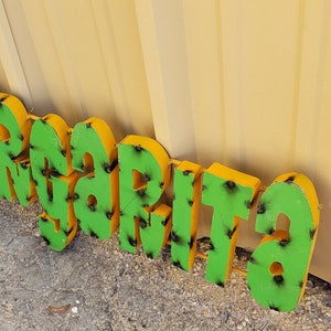 May include: A green and yellow metal sign that reads "Margarita". The sign is made of individual letters that are attached together.