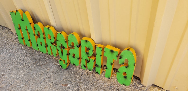 May include: A green and yellow metal sign that reads "Margarita". The sign is made of individual letters that are attached together.