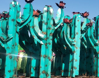 Könnte beinhalten: Eine Gruppe von tealfarbenen Metallkaktus-Skulpturen mit roten Metallblumen. Die Skulpturen sind in einer Reihe angeordnet und alle gleich groß. Sie sind aus Metall gefertigt und haben einen rustikalen Look.