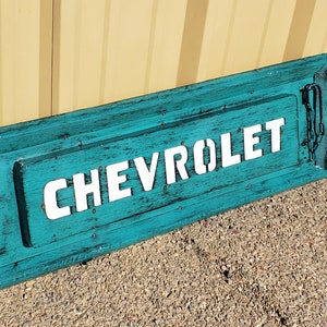 May include: A teal blue wooden sign with the word "CHEVROLET" in white lettering. The sign has decorative metal accents on each side.