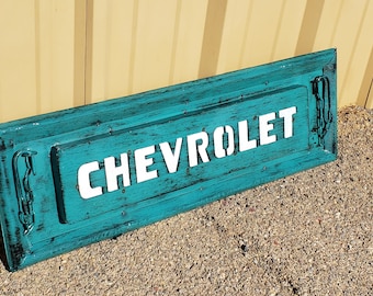 May include: A teal blue wooden sign with the word "CHEVROLET" in white lettering. The sign has decorative metal accents on each side.