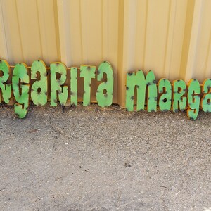 May include: Green metal letters spelling out "Margarita Margarita" on a gravel surface. The letters have a distressed look with some rust and are outlined in yellow.