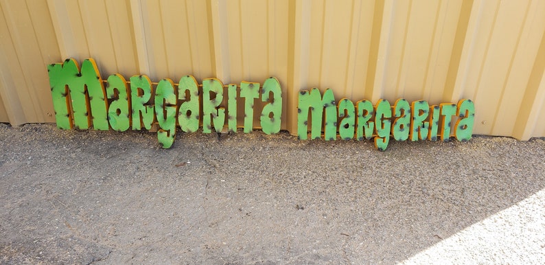 May include: Green metal letters spelling out "Margarita Margarita" on a gravel surface. The letters have a distressed look with some rust and are outlined in yellow.