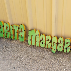 May include: Green and yellow metal letters spelling out "Margarita Margarita" on a gravel surface.