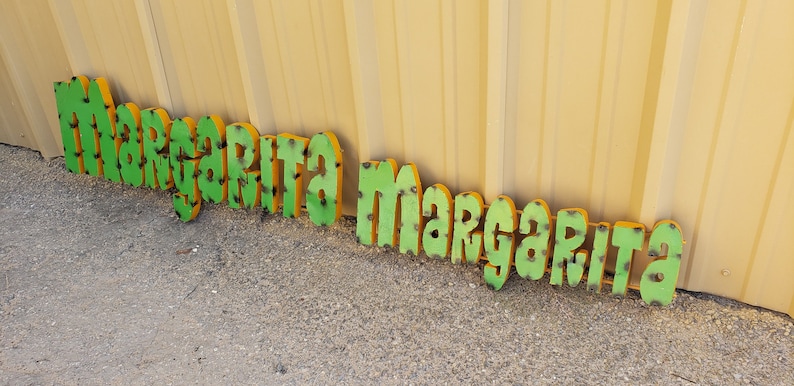 May include: Green and yellow metal letters spelling out "Margarita Margarita" on a gravel surface.