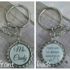 May include: A silver keyring with a school bus charm and a bottle top charm. The bottle top charm has the text "Ms. Cindy" on the front and "Thank you for always keeping us safe" on the back.