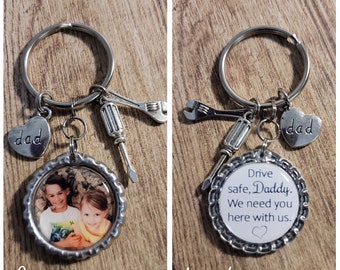 May include: A silver keychain featuring a photo pendant and charms. The front of the pendant displays two children. The back has the text "Drive safe, Daddy. We need you here with us." Charms include a heart with "dad" and miniature tools.