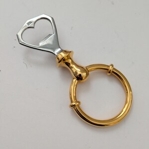 May include: A bottle opener with a heart-shaped top and a gold-colored handle and ring. The opener is made of silver-colored metal and gold-colored metal. The ring is designed for easy handling and storage.