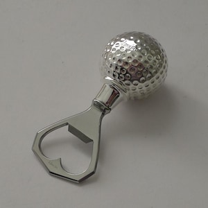 May include: A silver-toned metal bottle opener featuring a handle shaped like a golf ball with a dimpled texture. The word HERMES is engraved on the golf ball handle and on the lower edge of the opener mechanism, which has a heart-shaped cutout.