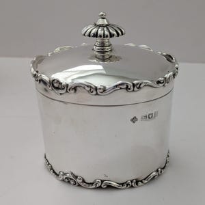 Puede incluir: Un recipiente cilíndrico plateado con tapa. La tapa tiene un pomo decorativo y un borde festoneado. El borde y la base del recipiente presentan adornos de volutas. La superficie es reflectante, lo que sugiere un acabado pulido.