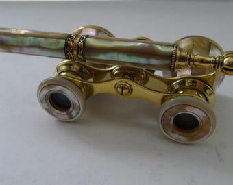 May include: Antique opera glasses with a gold-tone frame and iridescent mother-of-pearl accents. The central barrel is adorned with ornate gold detailing. The lenses are framed with mother-of-pearl, adding to the vintage aesthetic.