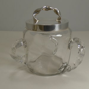 Puede incluir: Un tarro de cristal transparente con tapa y asa plateadas. El tarro tiene dos asas de cristal curvadas en los laterales. La tapa tiene un asa plateada retorcida. El tarro está vacío y se encuentra sobre una superficie blanca.