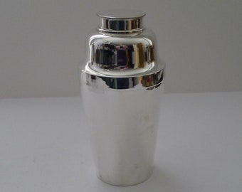 May include: A shiny, silver cocktail shaker with a rounded base and a cap. The shaker reflects the surrounding environment, creating a mirrored effect. The design is classic and elegant, suitable for mixing drinks.