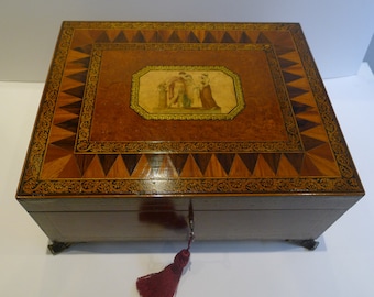 May include: A rectangular wooden box with a decorative inlaid top. The top features a central framed image of figures, surrounded by intricate geometric patterns in various wood tones. A key with a red tassel hangs from the lock.