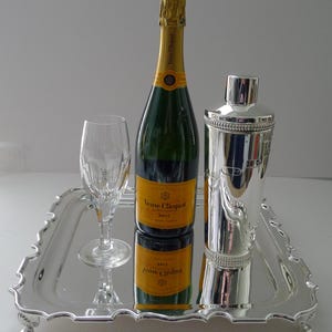 Könnte beinhalten: Ein silbernes Serviertablett mit einer Champagnerflasche, einem Cocktailshaker und einer Champagnerflöte. Die Champagnerflasche hat ein gelbes Etikett und Goldfolie. Der Cocktailshaker ist silberfarben und hat ein dekoratives Design.