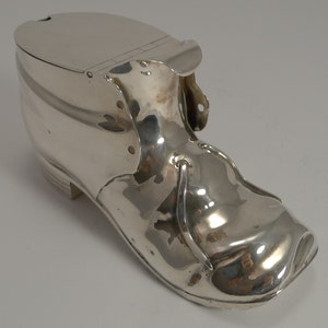 May include: A silver-colored shoe-shaped container with a hinged lid. The container has a smooth, reflective surface and is designed to resemble a boot. The shoe has a small heel and decorative rivets along the side.