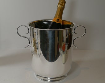 Puede incluir: Un cubo de champán plateado brillante con dos asas curvas. Dos botellas de champán con etiquetas doradas están dentro del cubo. El cubo tiene una base ancha y una superficie lisa y reflectante.