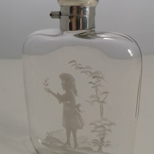 May include: Clear glass flask with a silver-colored, spherical stopper. The flask features an etched design of a young person holding a sprig, with stylized trees. The flask is rectangular with rounded edges.